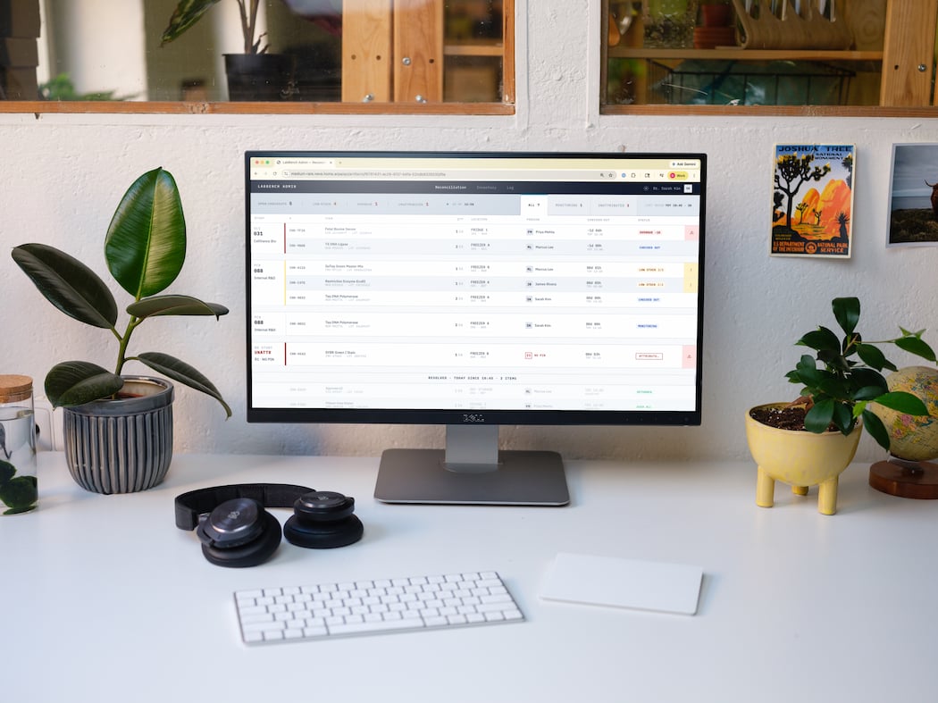 Desktop computer displaying an inventory management screen on a white desk with plants, headphones, keyboard, and mouse pad.