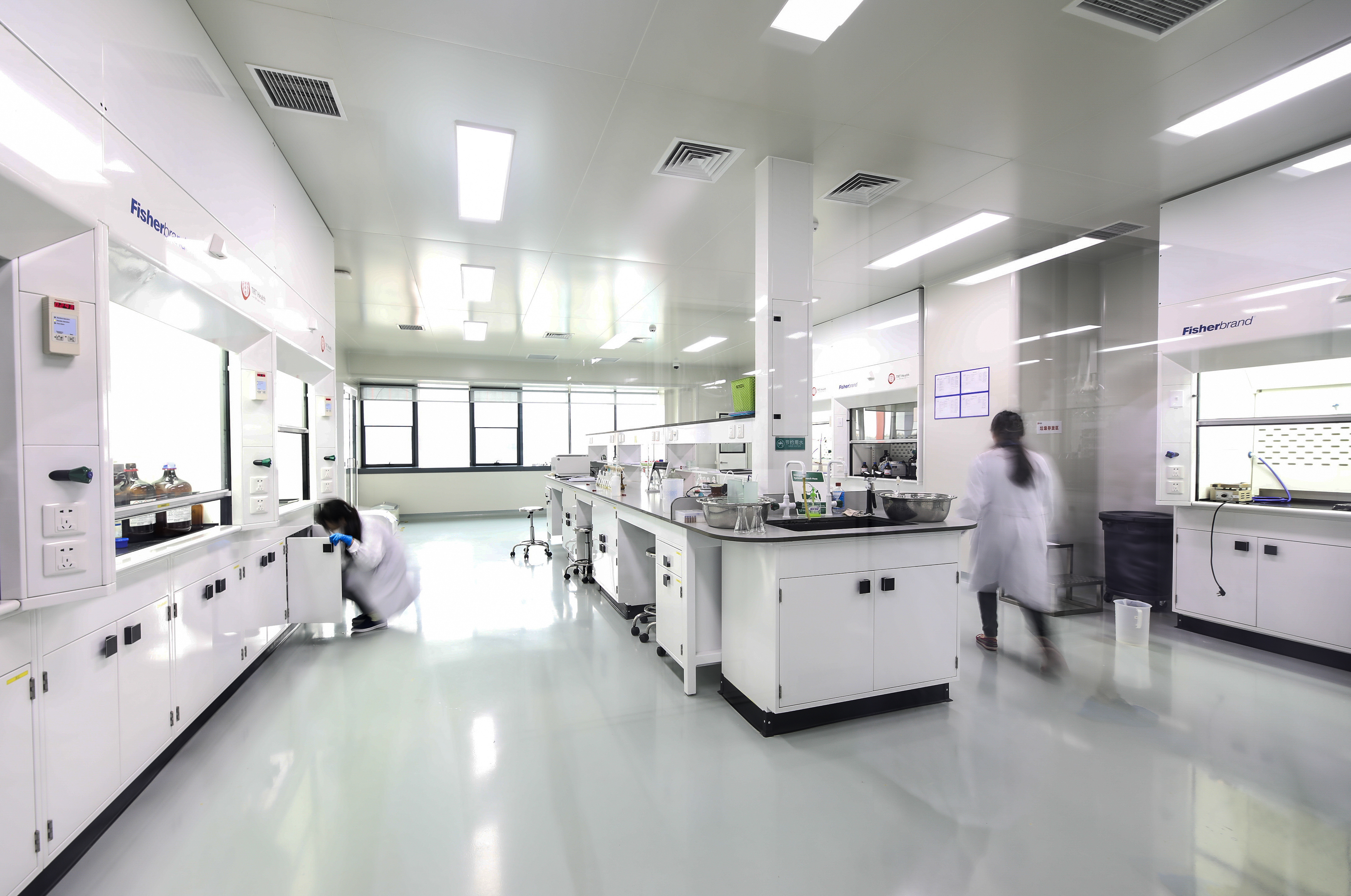 Bright modern laboratory with white cabinets, two scientists in lab coats working, and large windows.