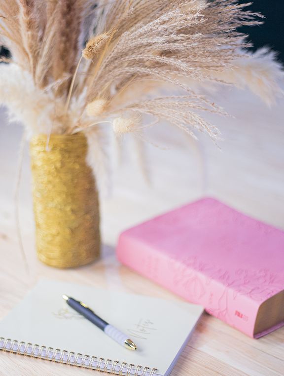 Close-up of dried grasses in a golden vase next to a pink bible and a notebook with a pen on a wooden surface.