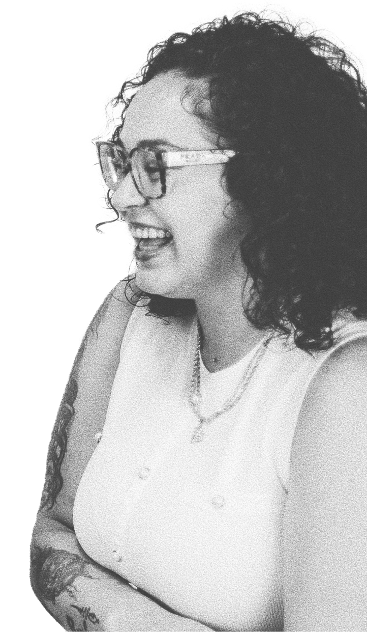 Smiling woman with curly hair, glasses, tattoos, and a sleeveless top, viewed in black and white.