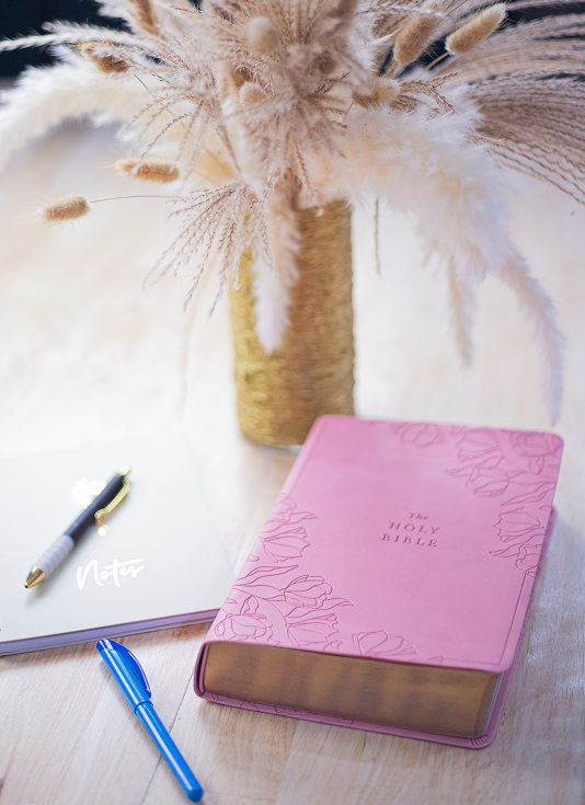 Pink Holy Bible with floral design beside a notebook labeled Notes and two pens on a wooden table.