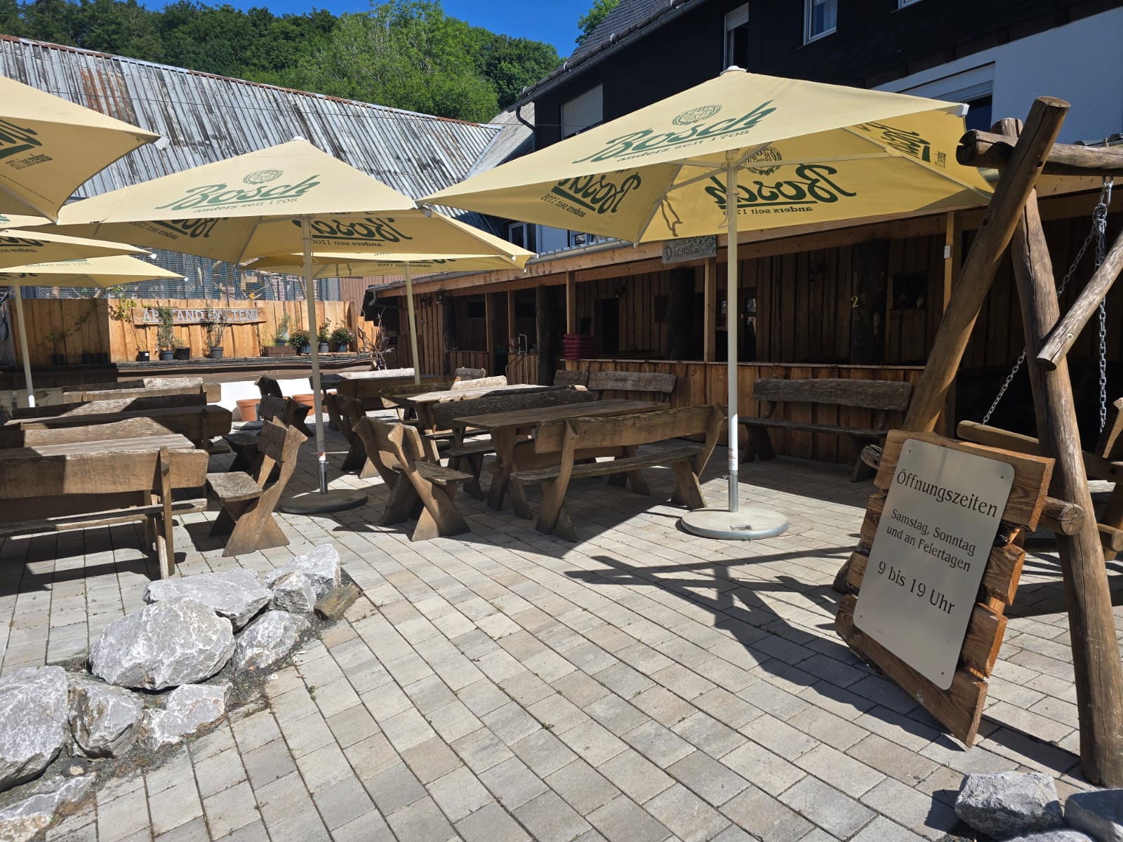 Außenbereich eines Biergartens mit Holztischen, Bänken und Sonnenschirmen unter blauem Himmel.