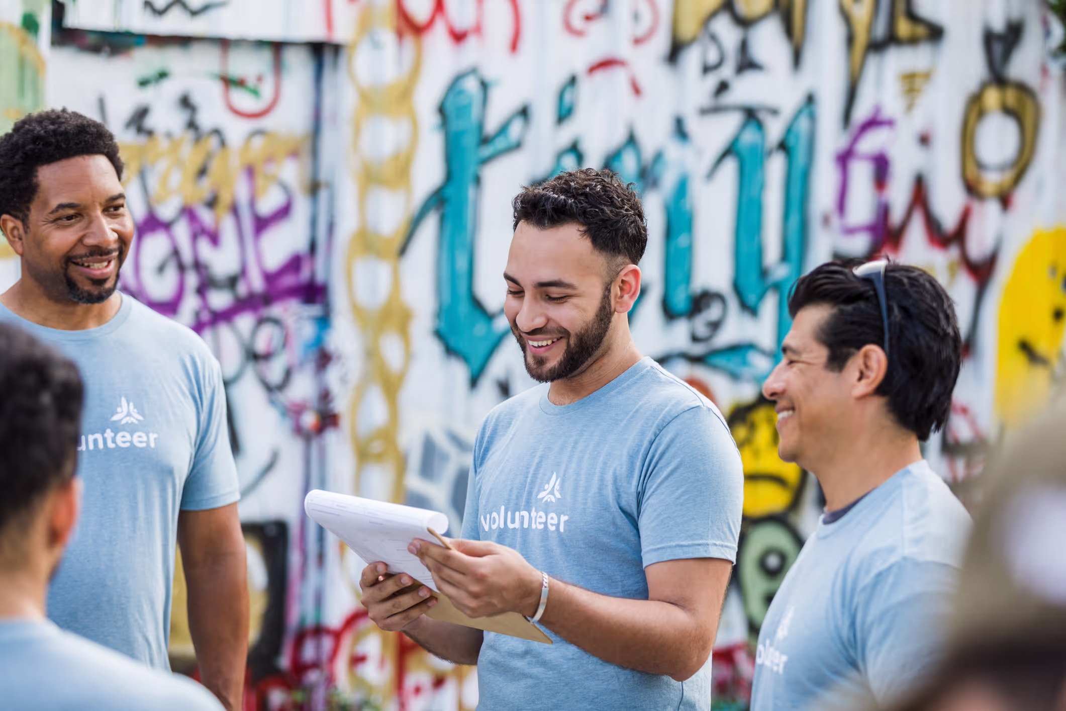 Group of smiling volunteers wearing light blue shirts standing and talking near a graffiti-covered wall.