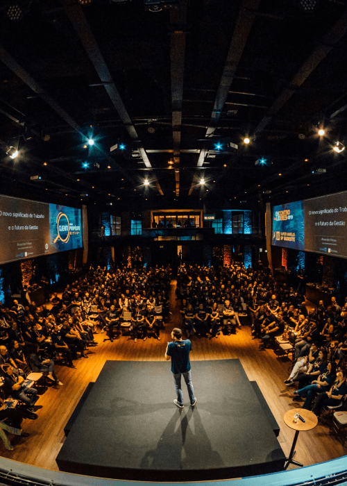 Speaker standing on a stage in a large auditorium addressing a seated audience with two large screens displaying presentation content.