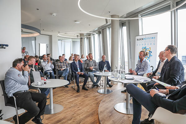Panel discussion at a blockchain conference with speakers seated in front and audience listening in a modern room with large windows.