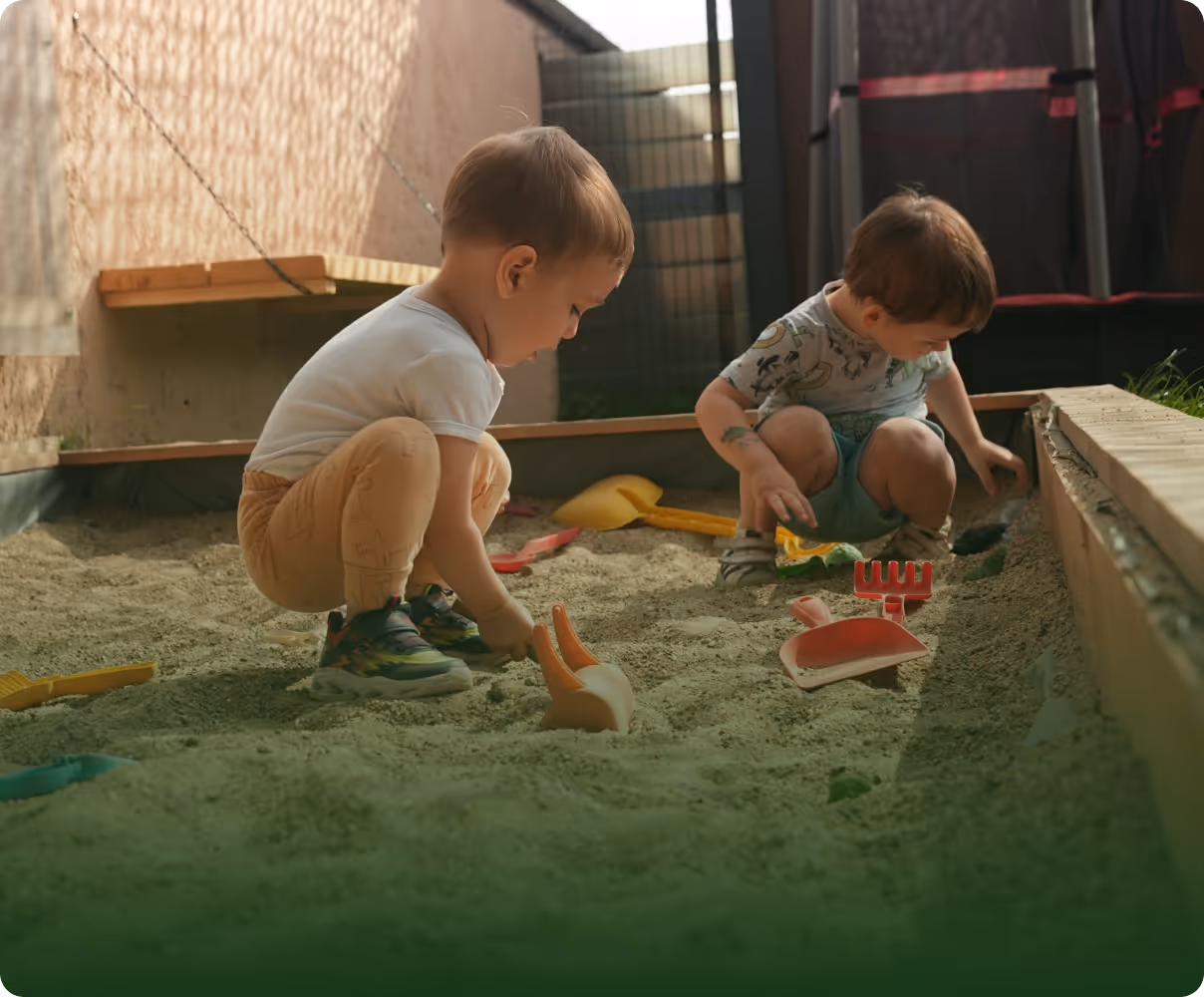 Two young children playing with plastic shovels and rakes in a sandbox outdoors.