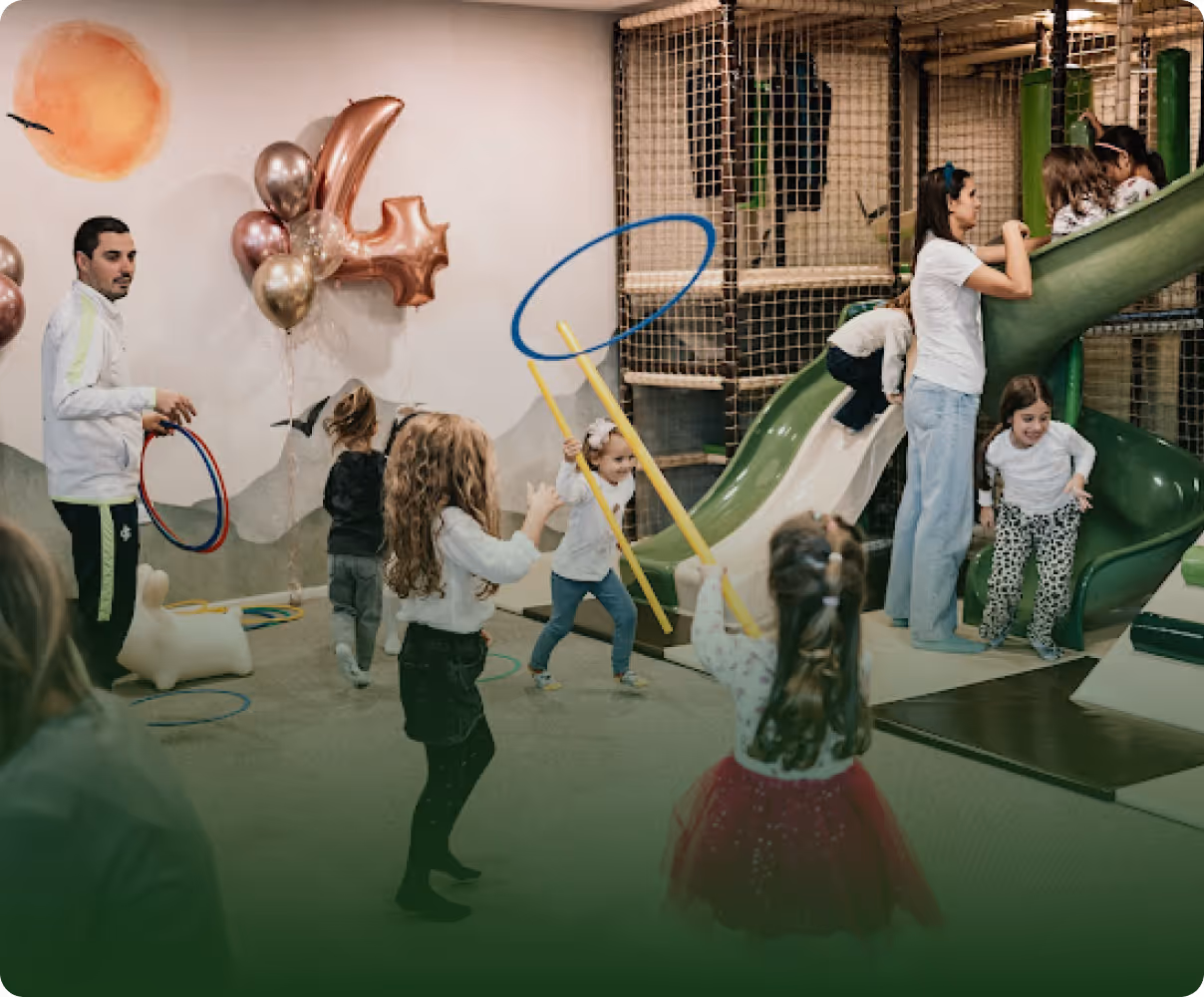 Children playing indoors with an adult supervising near a slide and balloons shaped as the number 4.