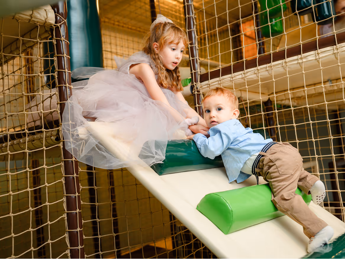 Young girl in a light pink dress helps a toddler boy climb up a padded play structure inside an indoor playground.