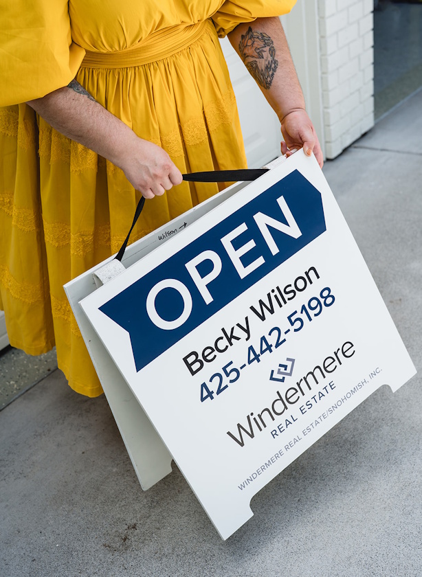 A person in a yellow dress holds an A‑frame open house sign with real estate branding.