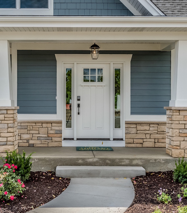 A modern house entrance with a white door, blue‑gray siding, stone accents, and landscaped flower beds.
