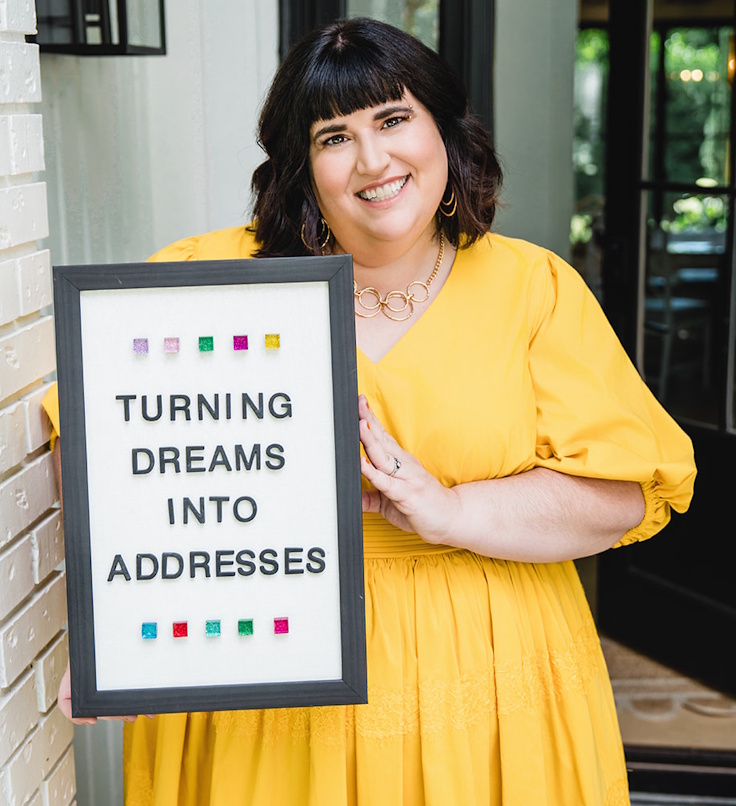 A person in a yellow dress holds a framed sign that reads “Turning Dreams Into Addresses.”