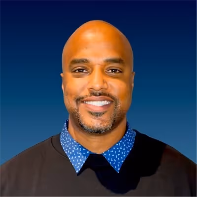 Smiling bald man with trimmed beard wearing a black sweater over a blue patterned shirt against a blue background.