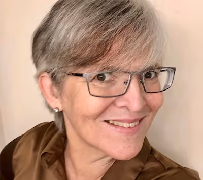 Smiling woman with short gray hair and glasses wearing a brown shirt against a beige background.