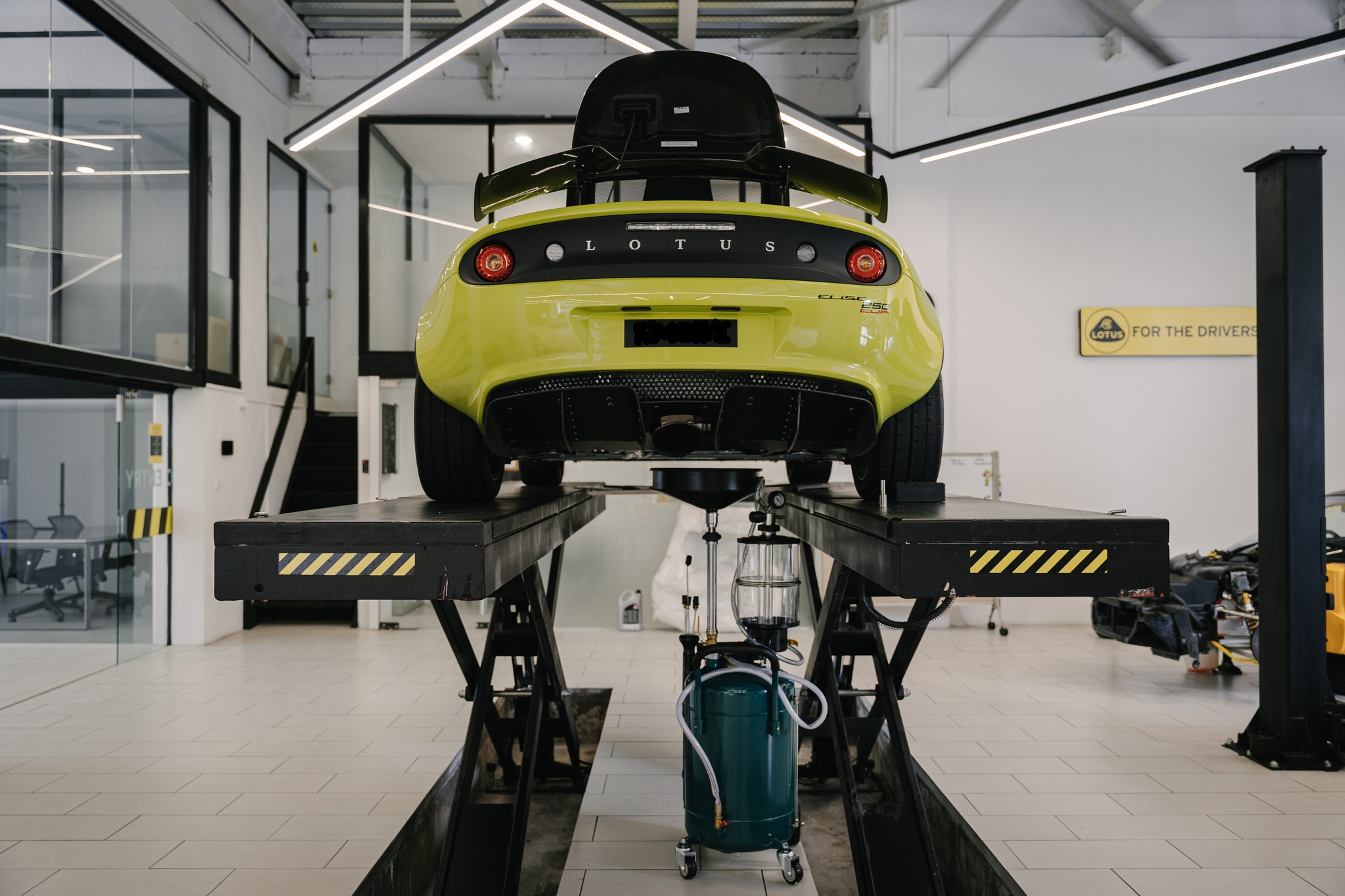Yellow Lotus Elise 250 cup sports car on a black hydraulic lift in a clean, modern car service garage.