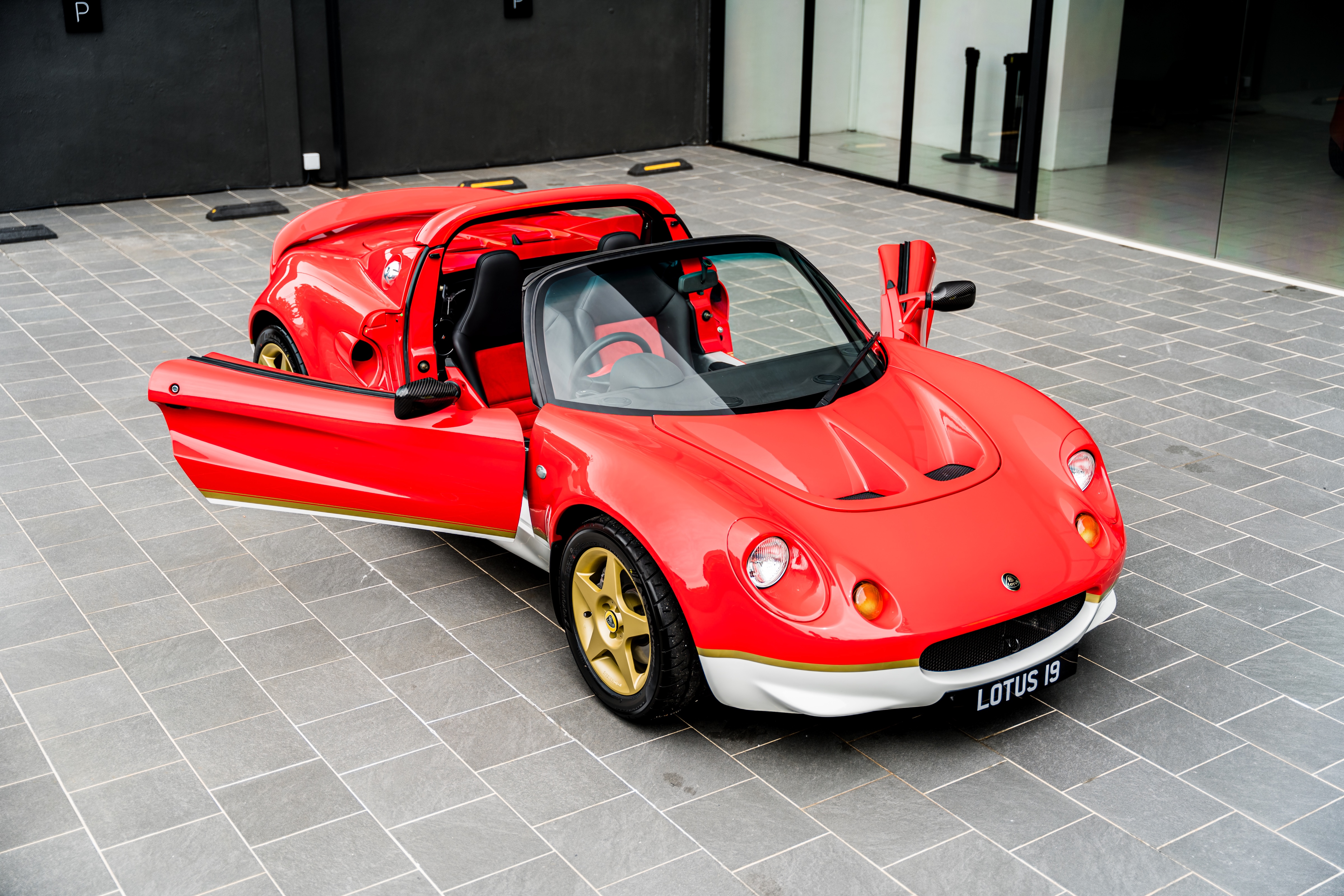 Red Lotus sports car with open doors and convertible roof parked on gray tiles.