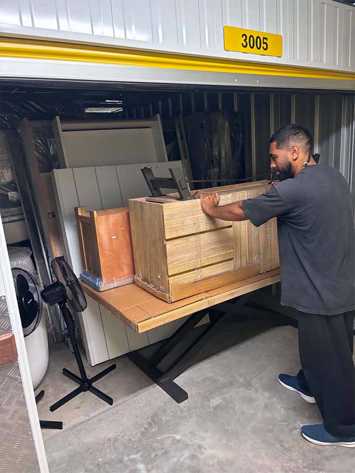 Man pushing wooden furniture on a table inside a storage unit numbered 3005.
