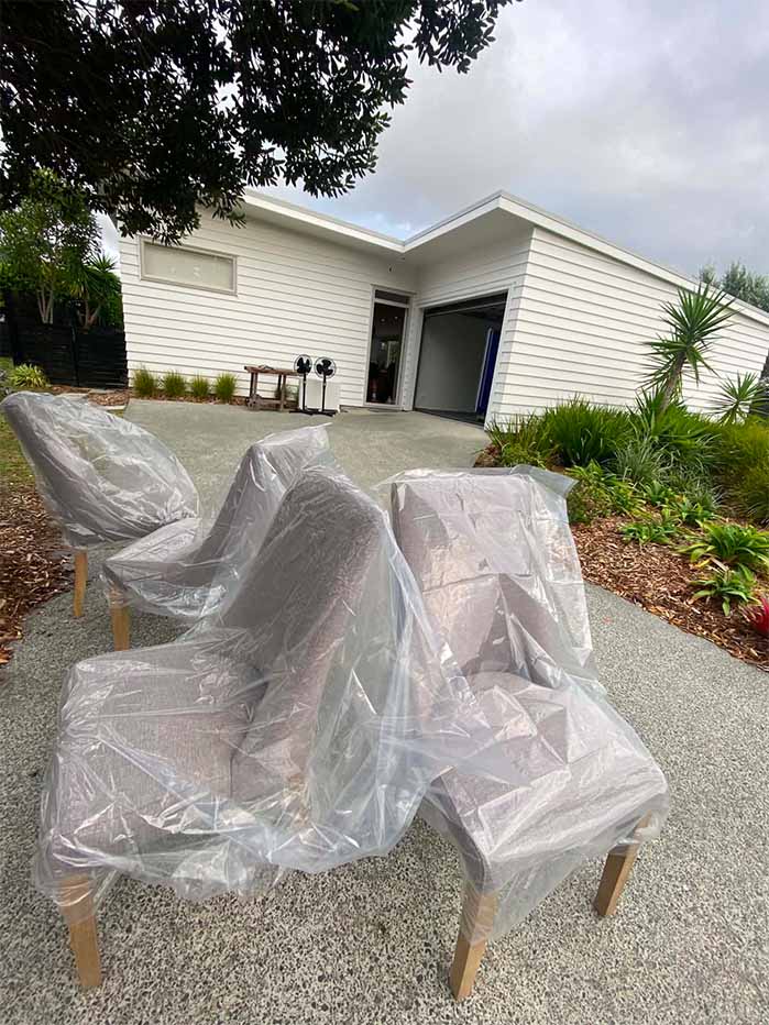 Four cushioned chairs wrapped in plastic on a paved driveway outside a modern white house.
