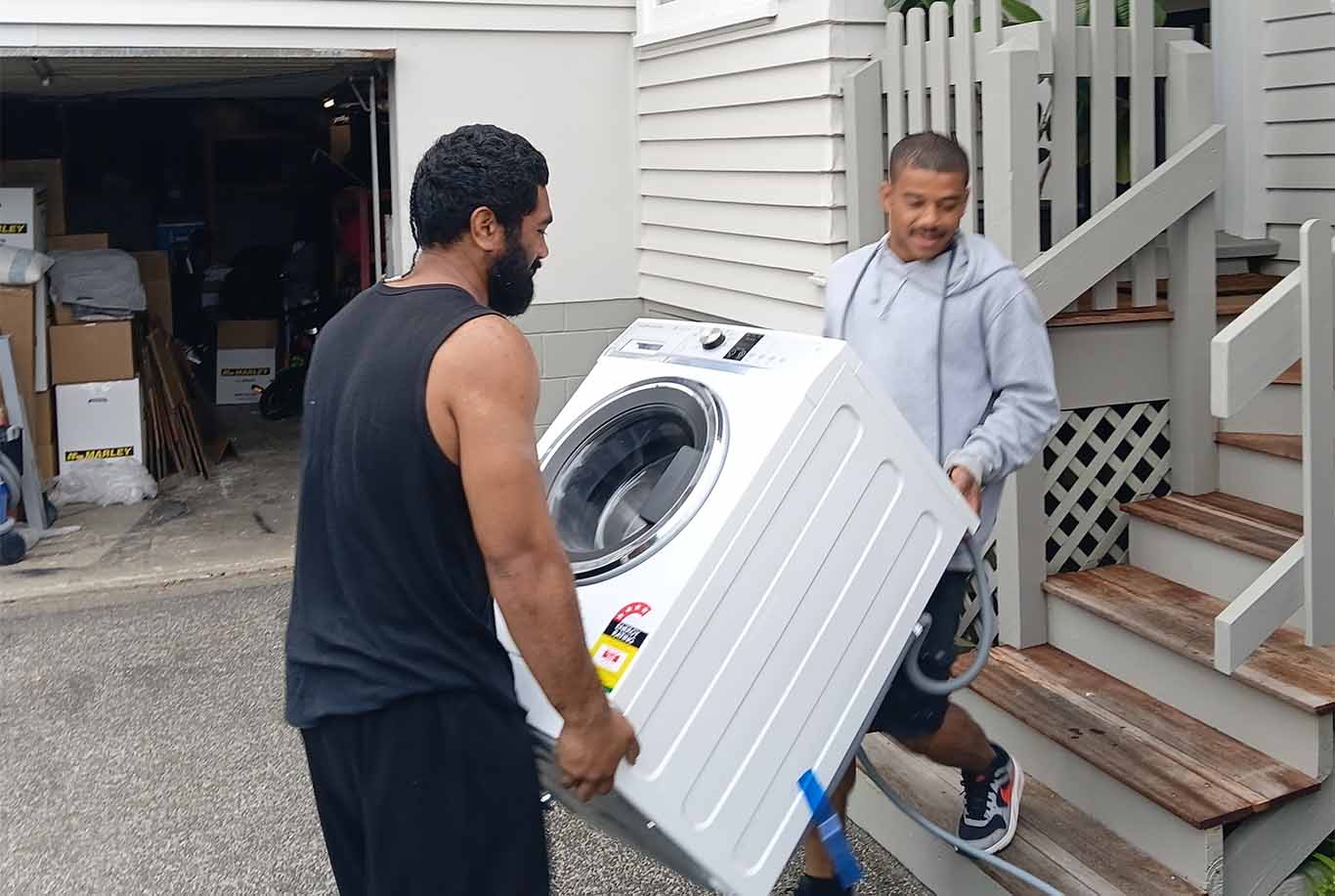 Two men carrying a washing machine up wooden stairs outside a house near a garage.