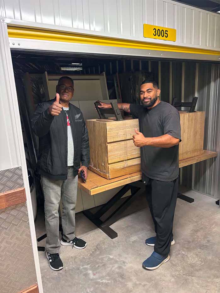 Two men giving thumbs up in a storage unit beside stacked wooden furniture pieces.
