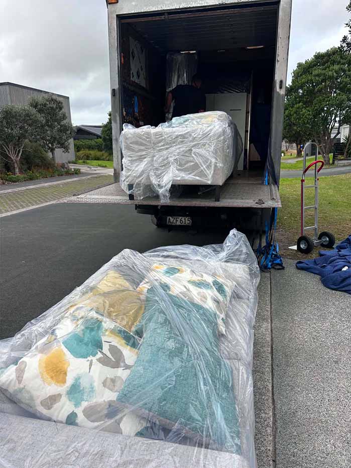 Wrapped couch cushions on the ground and wrapped sofa inside a moving truck with a dolly nearby.