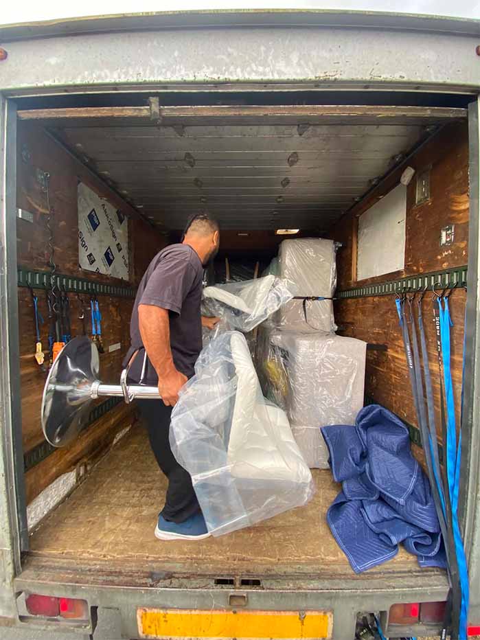 Man loading a plastic-wrapped chair and boxes inside a moving truck with straps and moving blankets.