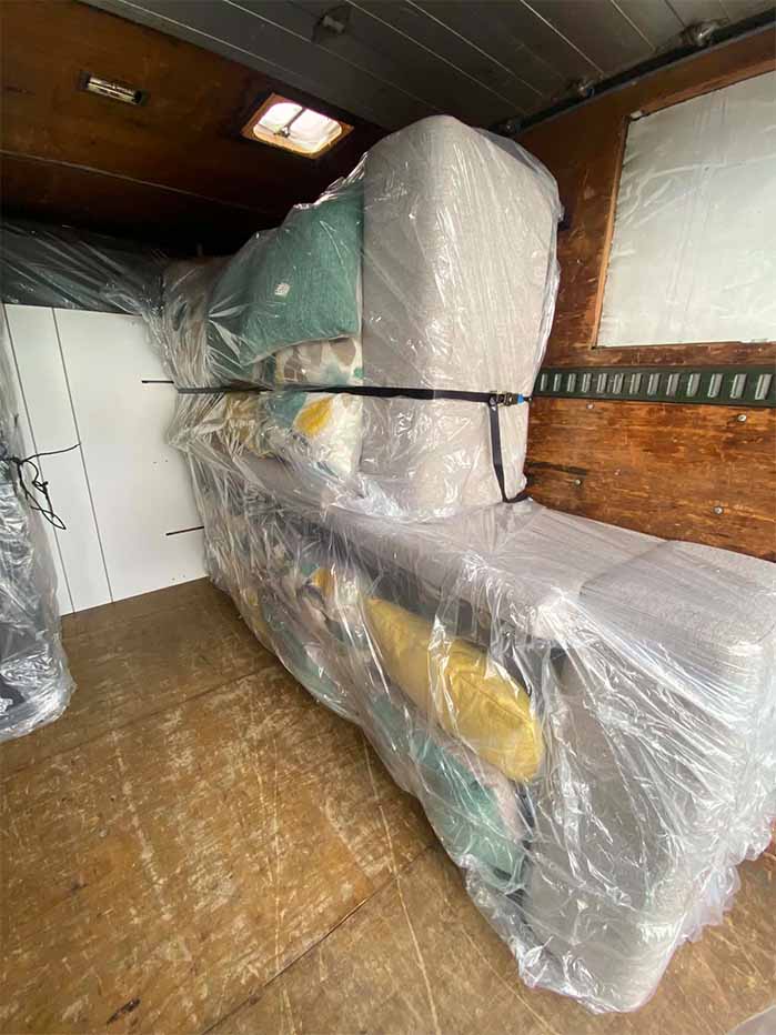 Furniture wrapped in plastic and secured with a strap in a wooden moving truck interior.