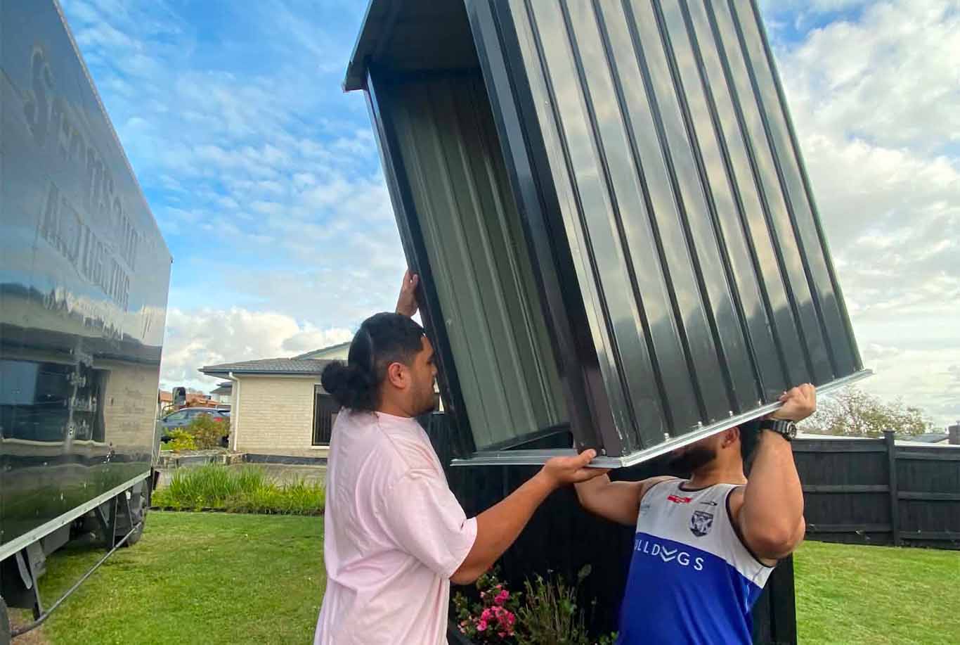 Two men outdoors lifting a large black metal shed panel in a grassy backyard.