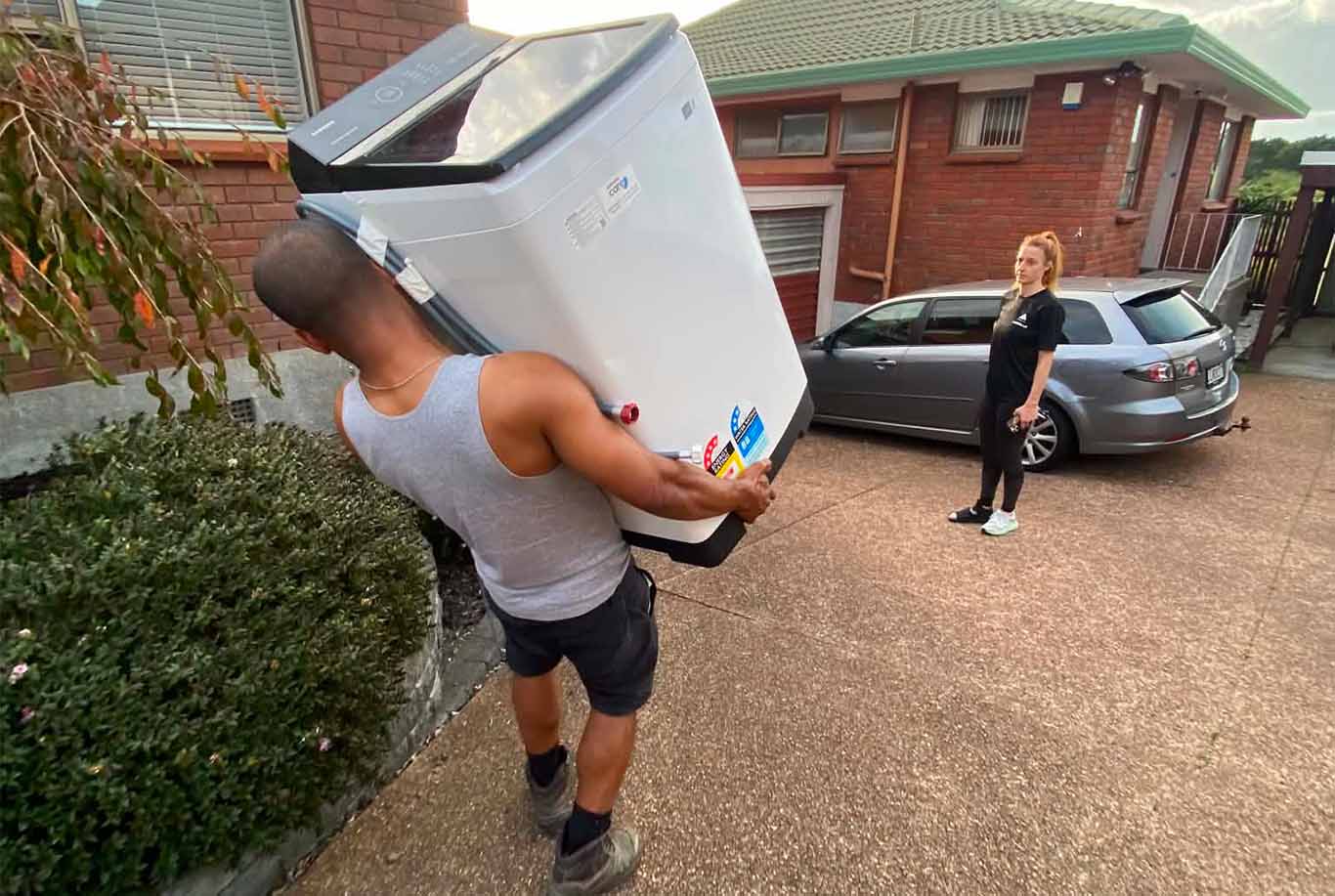 Man carrying a washing machine outdoors while a woman watches near a parked gray car by a brick house.