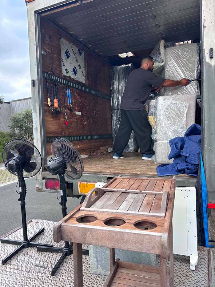 Man securing wrapped furniture inside a moving truck with fans and wooden cart nearby.
