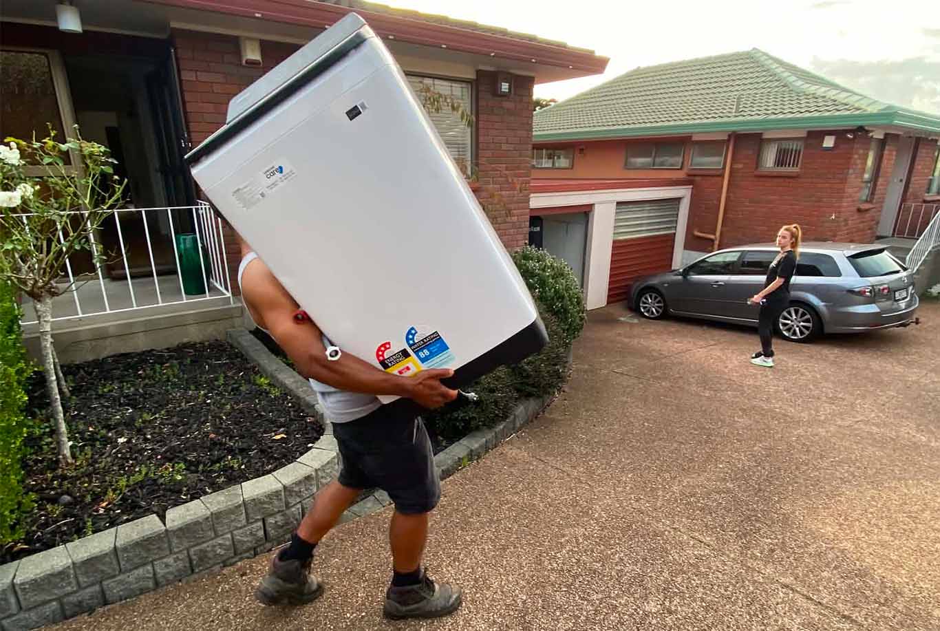 Person carrying a large white appliance outside a brick house with a woman standing near a parked car.