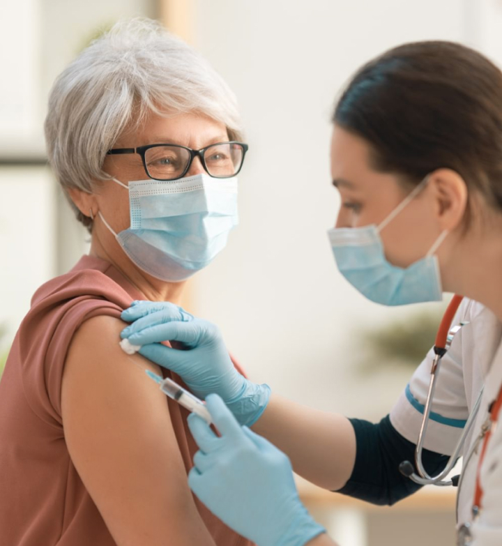 Healthcare professional wearing a mask and gloves administering a vaccine to an older woman wearing glasses and a mask.