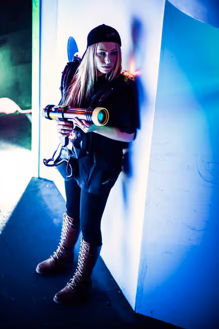 Young woman in black holds laser tag gun, leaning against a wall under blue and purple lights.