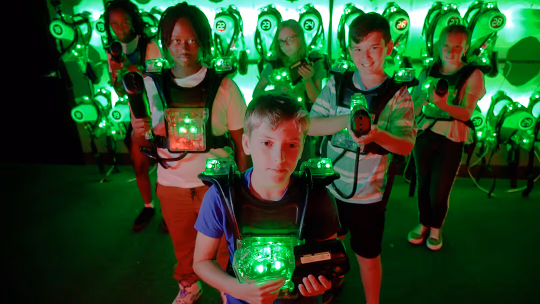 Five children wearing glowing green laser tag vests and holding laser guns in a game arena.