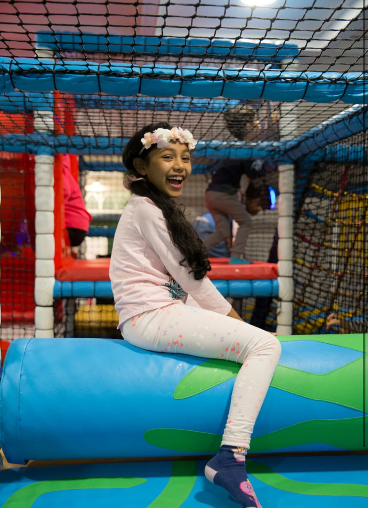 Smiling girl wearing floral headband sitting on blue padded play structure inside play area netting.