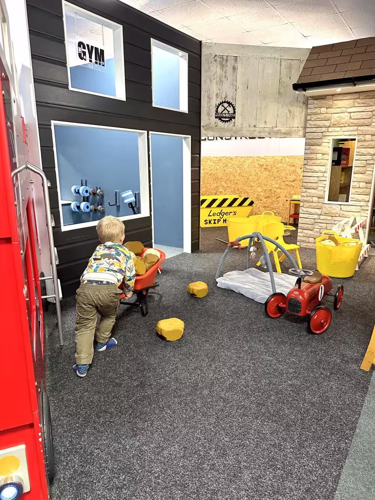 Child playing with toy wheelbarrow and blocks in an indoor play area with playhouse and toy car.