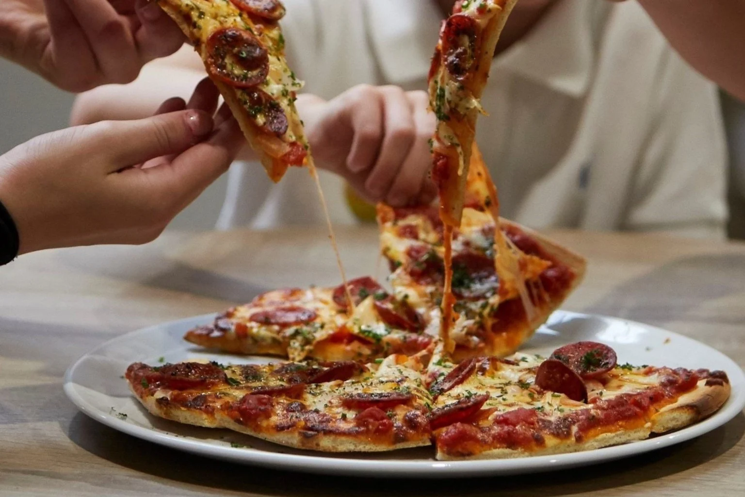 Two hands each holding a pepperoni pizza slice with melted cheese stretching from the pizza on a white plate.