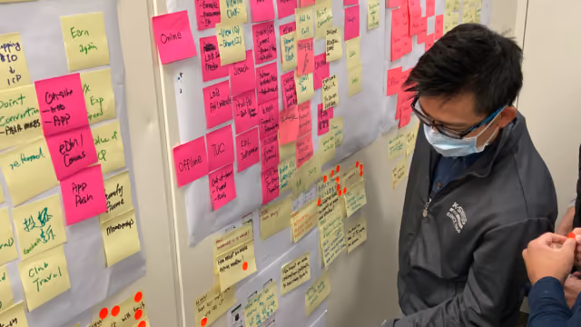 Person wearing glasses and mask looks at a wall covered with pink and yellow sticky notes with writing.