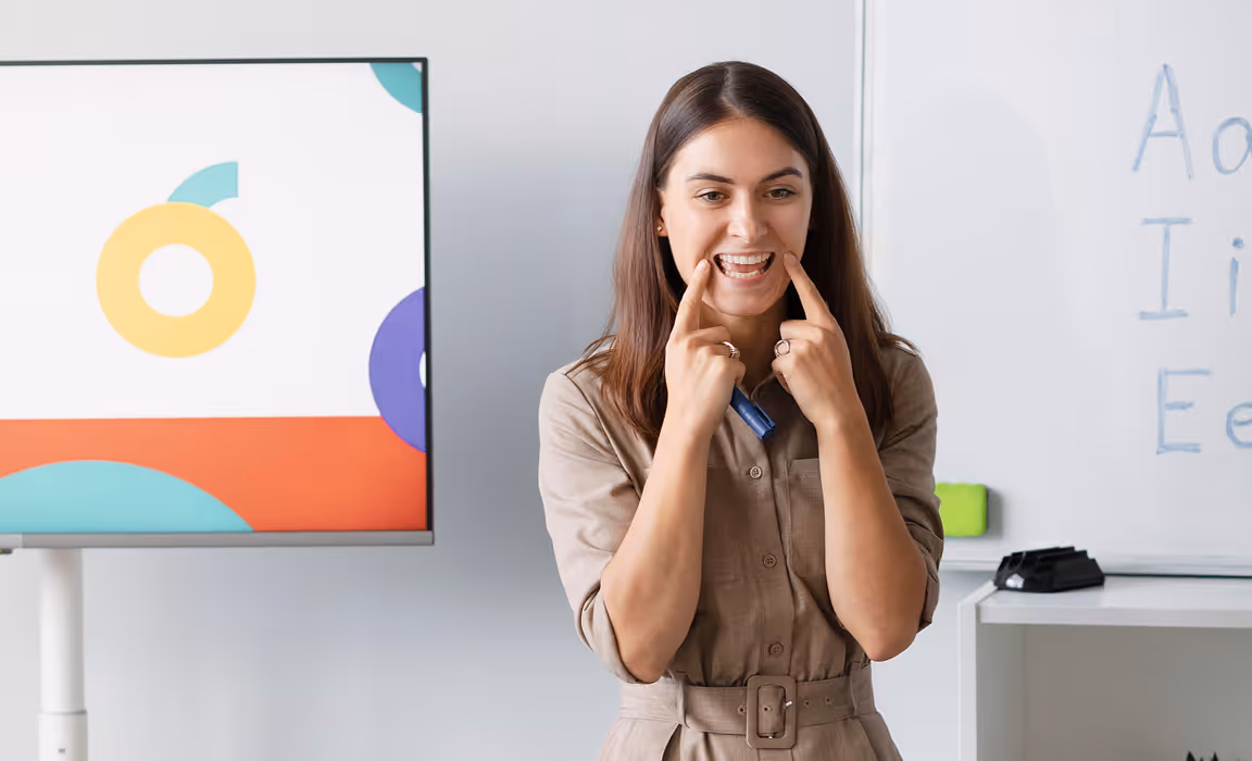 Une femme montrant ses dents avec les doigts dans une salle de classe avec un tableau et un écran coloré.