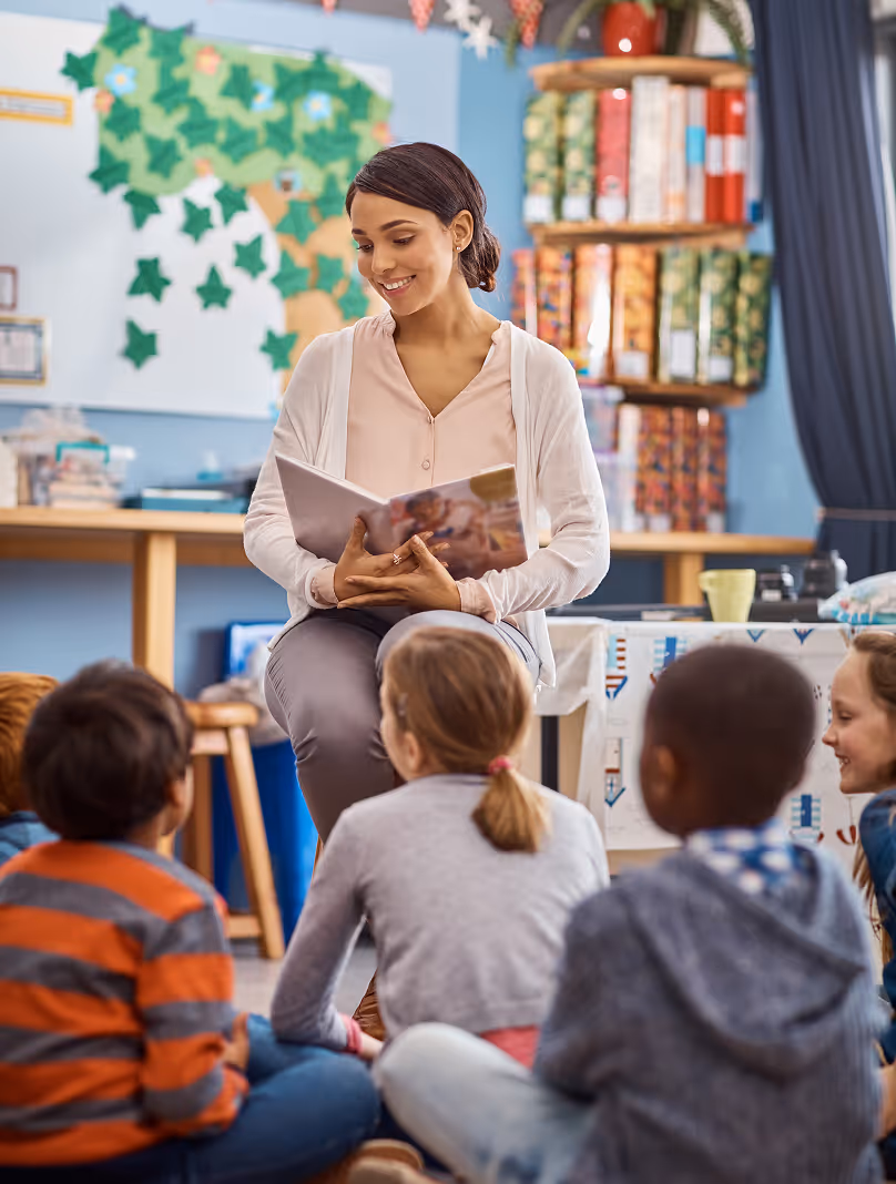Une enseignante souriante lisant un livre à un groupe d'enfants assis par terre en classe.
