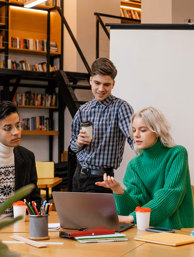Trois jeunes discutant devant un ordinateur portable dans une bibliothèque au style moderne.