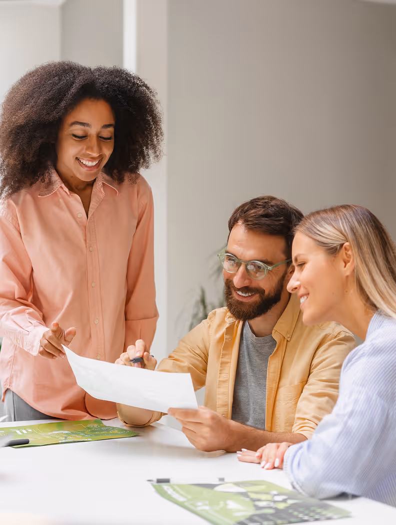 Trois collègues souriants discutent autour d'une table avec des documents de travail verts.