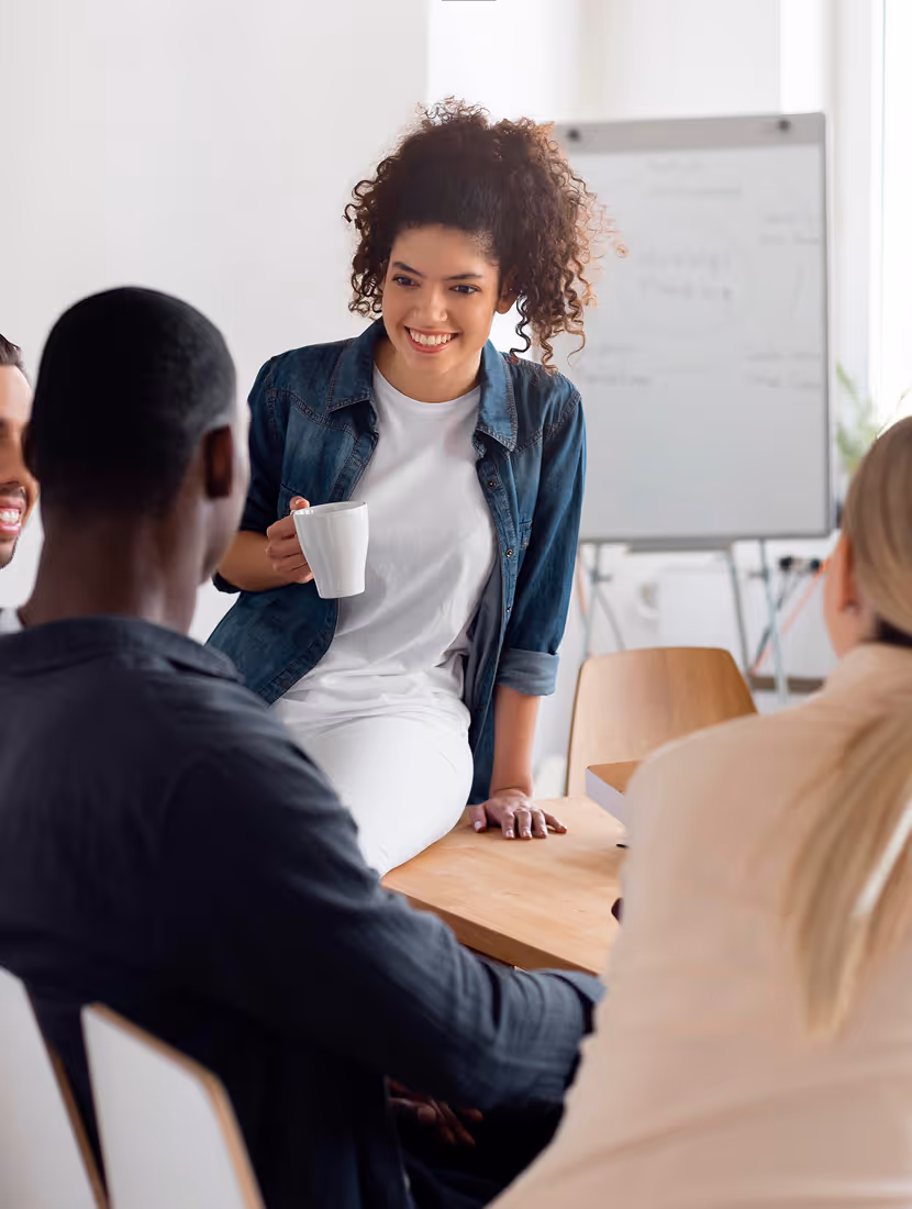 Jeune femme souriante avec tasse, discutant avec des collègues lors d'une réunion informelle.