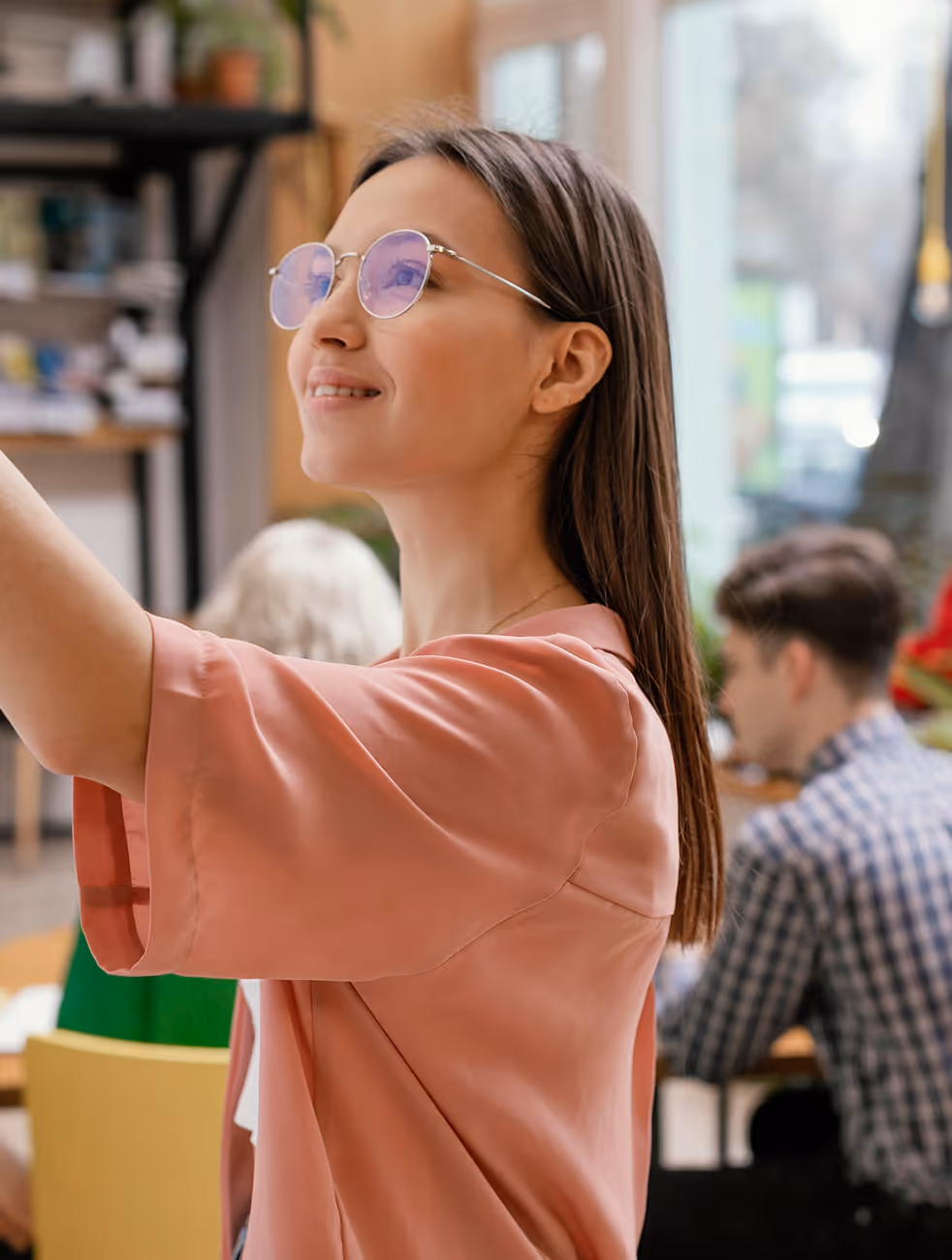 Femme portant des lunettes souriant et levant la main dans une salle de classe moderne.