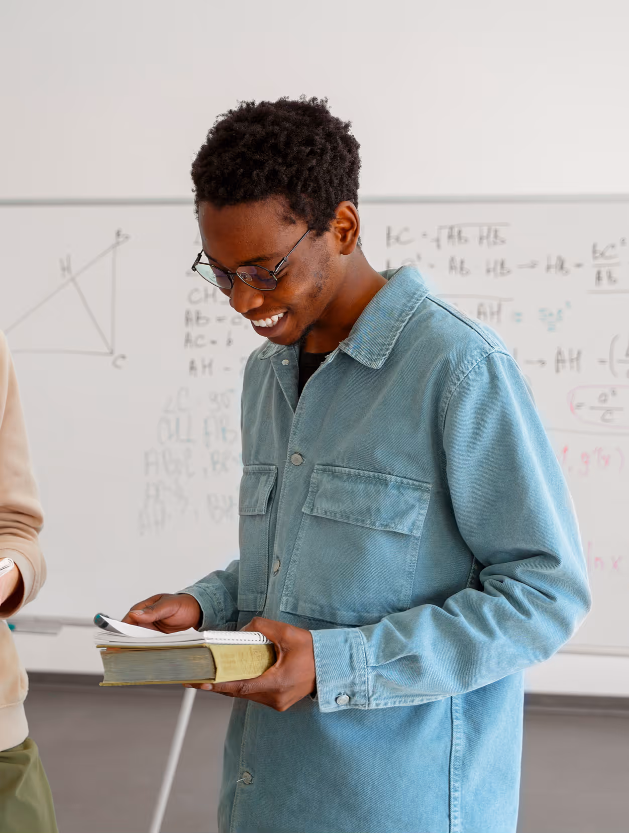 Jeune homme souriant avec des lunettes et une veste en jean tenant un livre devant un tableau blanc.