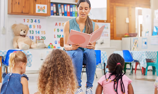Une enseignante lit un livre rose à trois enfants assis dans une salle de classe colorée.