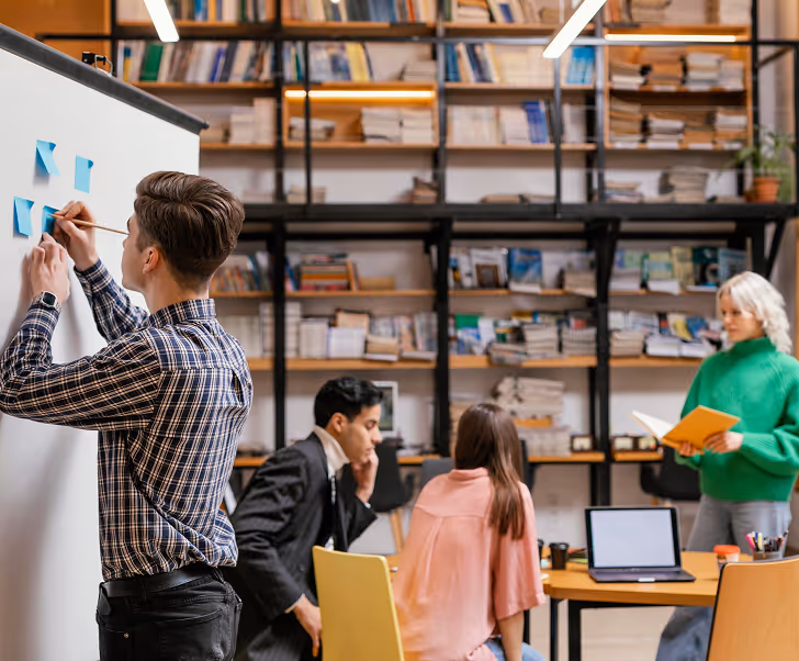 Un homme place des notes sur un tableau blanc devant trois collègues dans un bureau avec une grande bibliothèque.