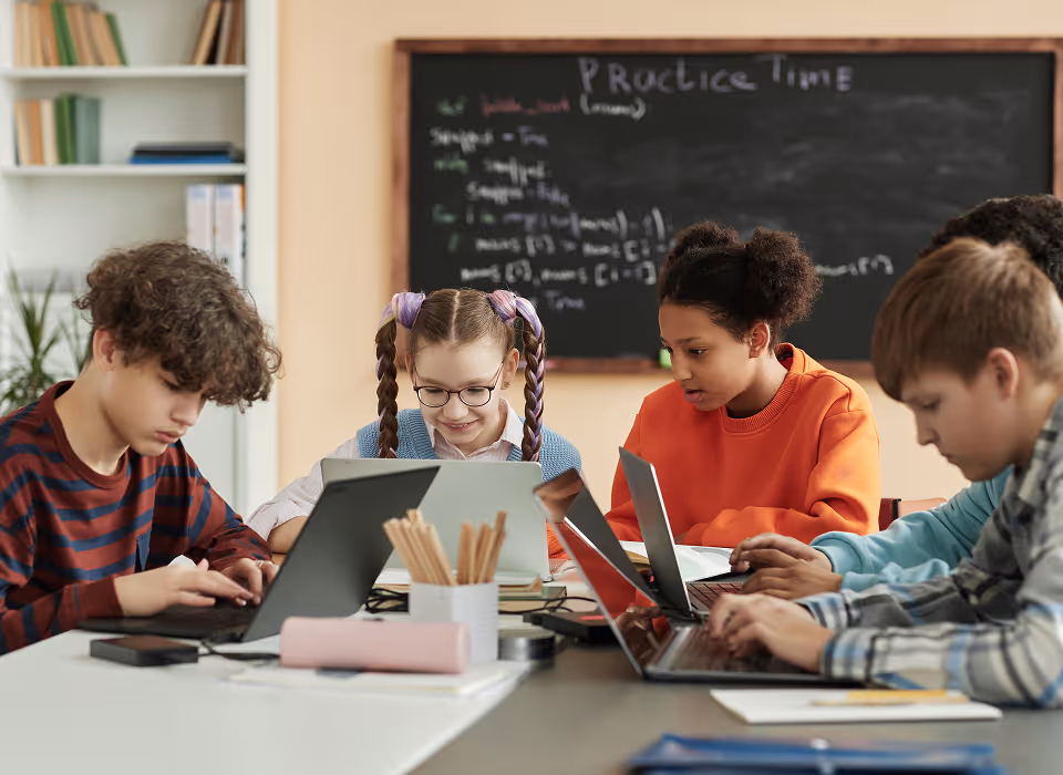 Quatre élèves concentrés travaillant sur des ordinateurs portables et une tablette en classe.