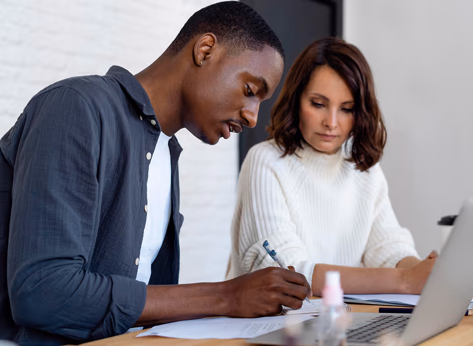 Un homme écrit tandis qu'une femme en pull blanc regarde, assis à une table avec un ordinateur portable.