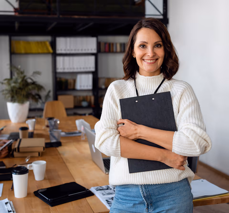 Femme souriante en pull blanc tenant un classeur noir dans un bureau moderne.