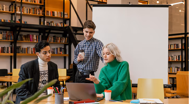 Trois jeunes adultes discutent autour d’un ordinateur portable dans une bibliothèque moderne.